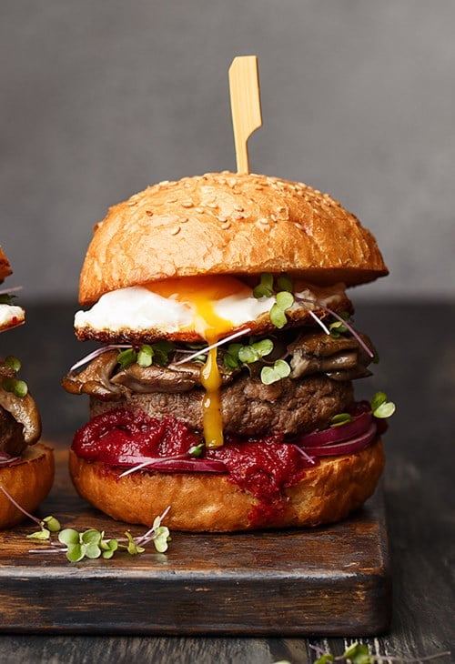 A plated burger stacked tall with muchsrooms, egg, and onions held together with a toothpick
