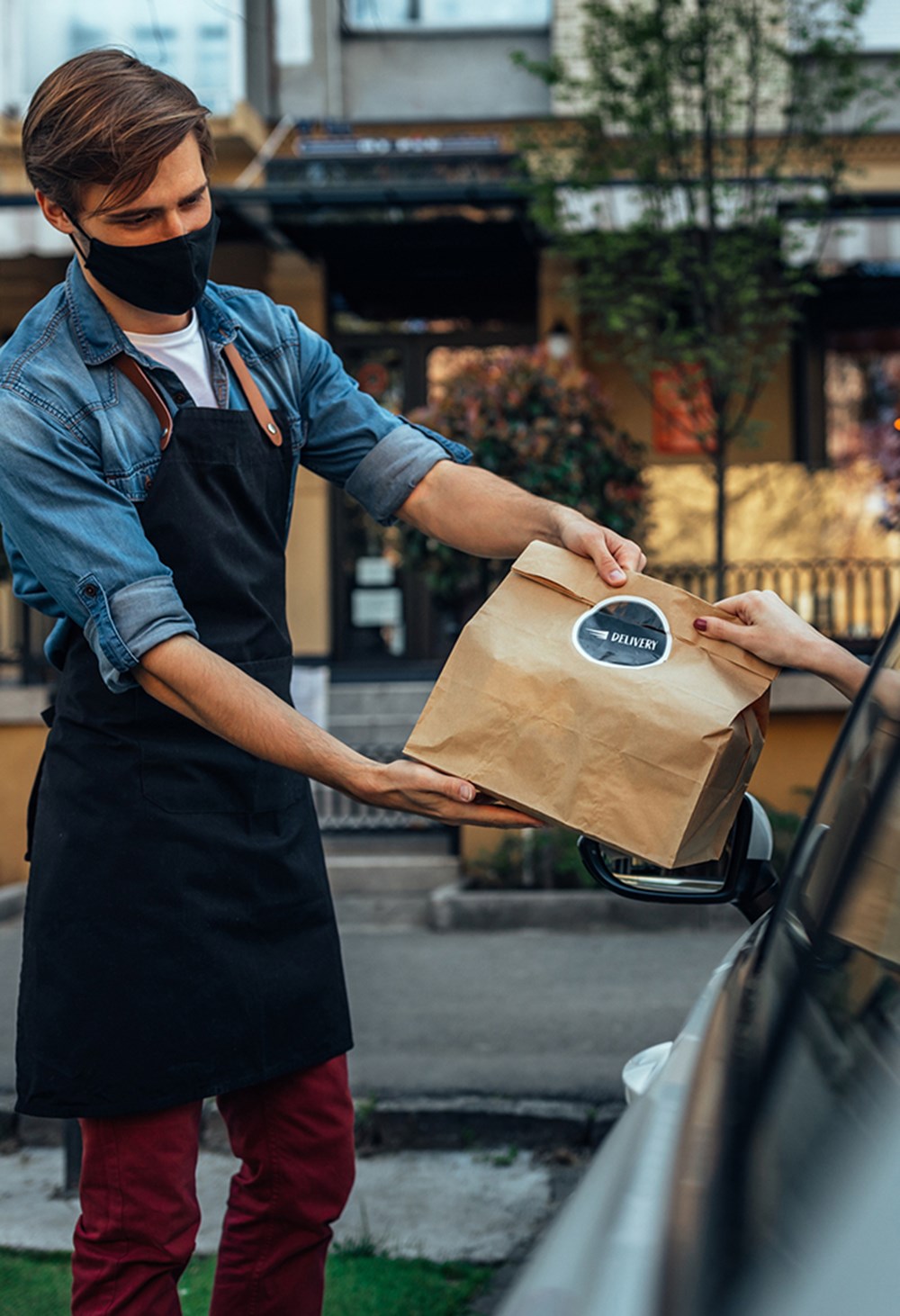 A masked restaurant employee handing a sealed curbside order to a customer in a parked car.