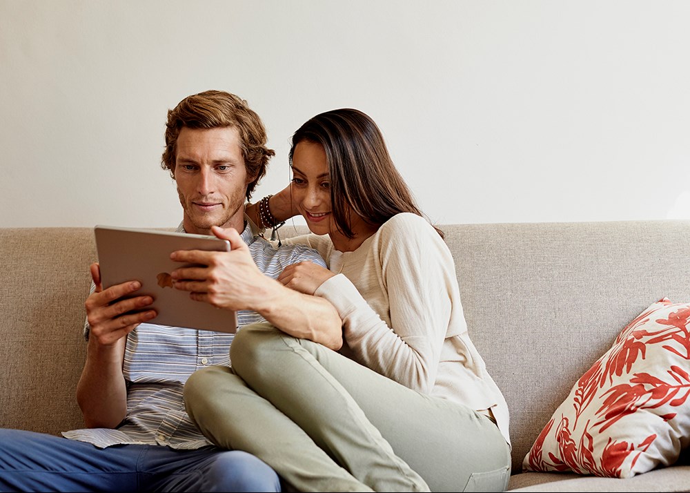 A couple sitting on a couch while looking at a tablet.