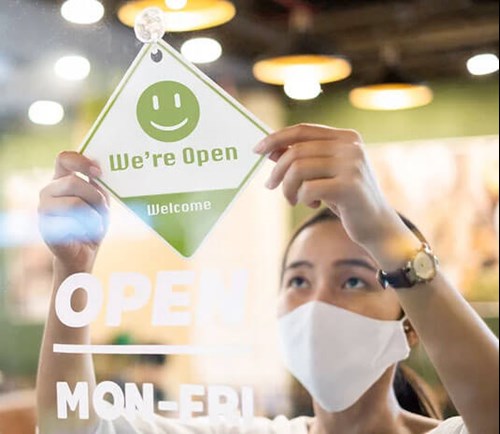 A woman wearing a mask hanging a sign that reads we're open.