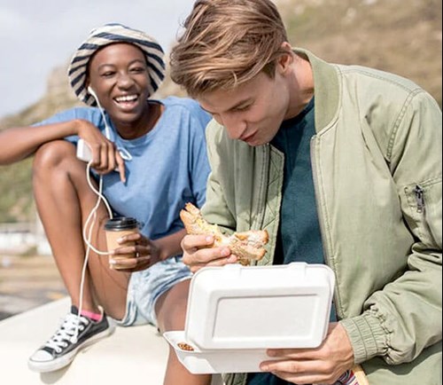 A man eating a sandwich from a to-go container while his friend laughs.