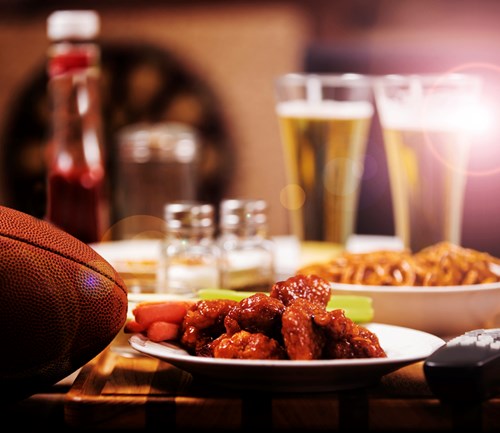 Buffalo wings, pretzels, beer, and a football on a table next to a remote control.
