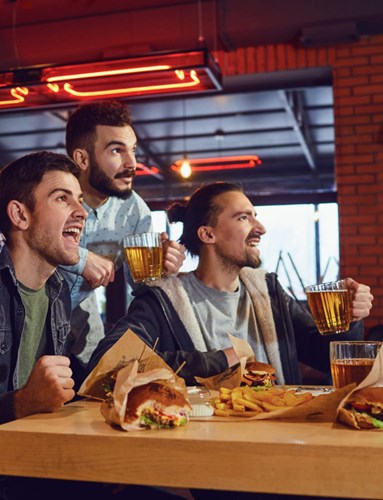 Three friends holding beers and watching football in a restaurant with sandwiches and french fries on the table.